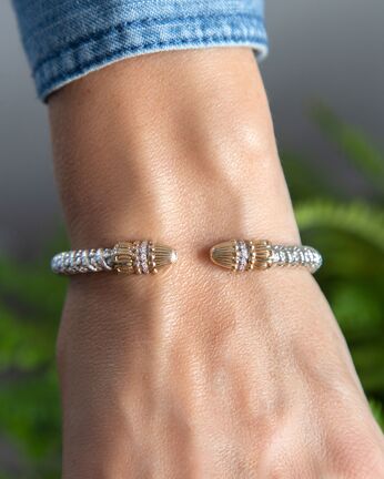 Close-up of a wrist wearing two gold bracelets with a blurred natural background