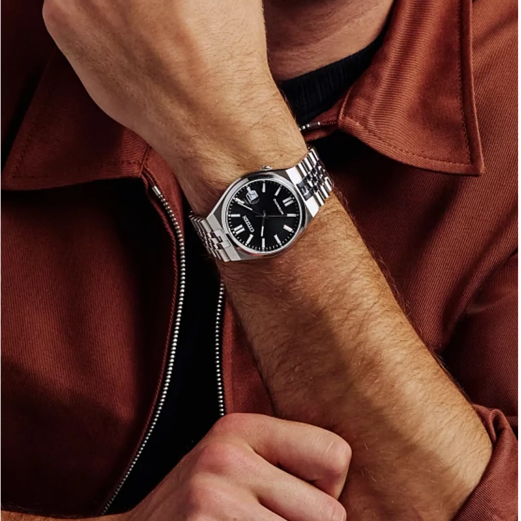 Close-up of a wrist wearing a silver watch with a black face, against a brown jacket background.