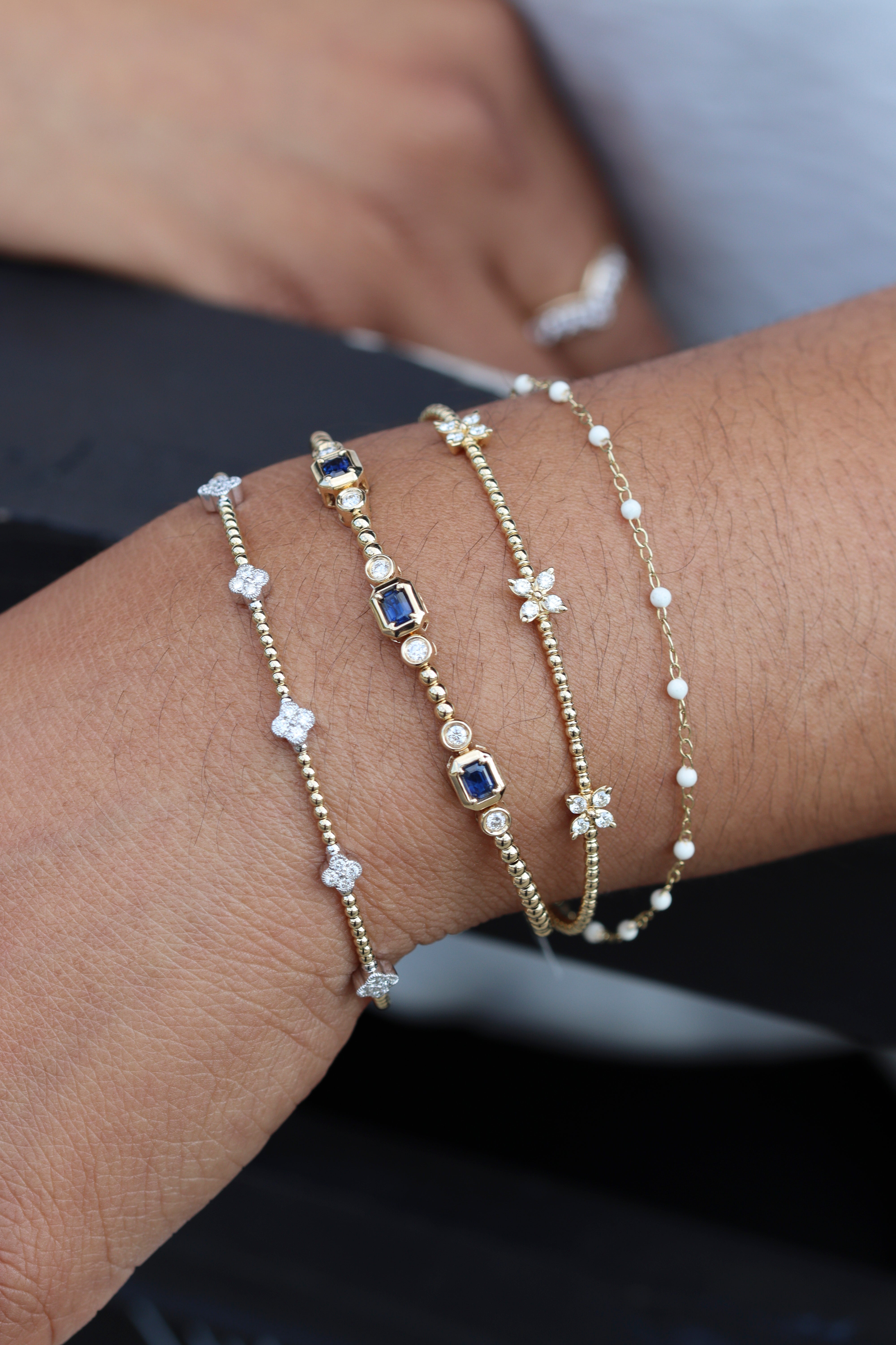 A close-up of a person's wrist wearing four delicate gold bracelets. The bracelets include clear crystal floral and star motifs, small white pearl beads, and two square-cut dark blue gemstone accents.
