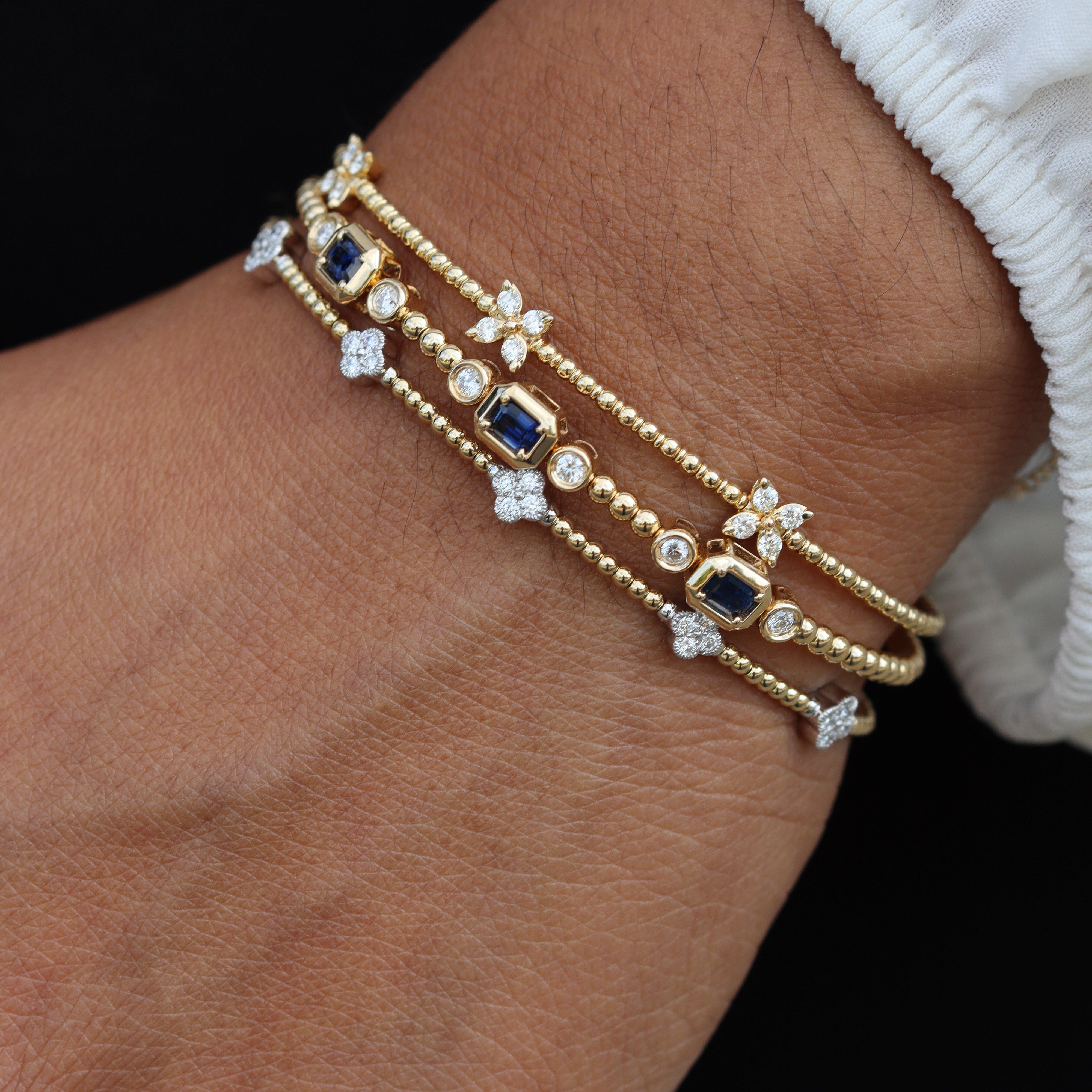 A close-up of a person's wrist wearing a stack of three delicate gold bracelets. The bracelets feature small gold beads, clear pavé-set round stones, and rectangular blue gemstone accents.