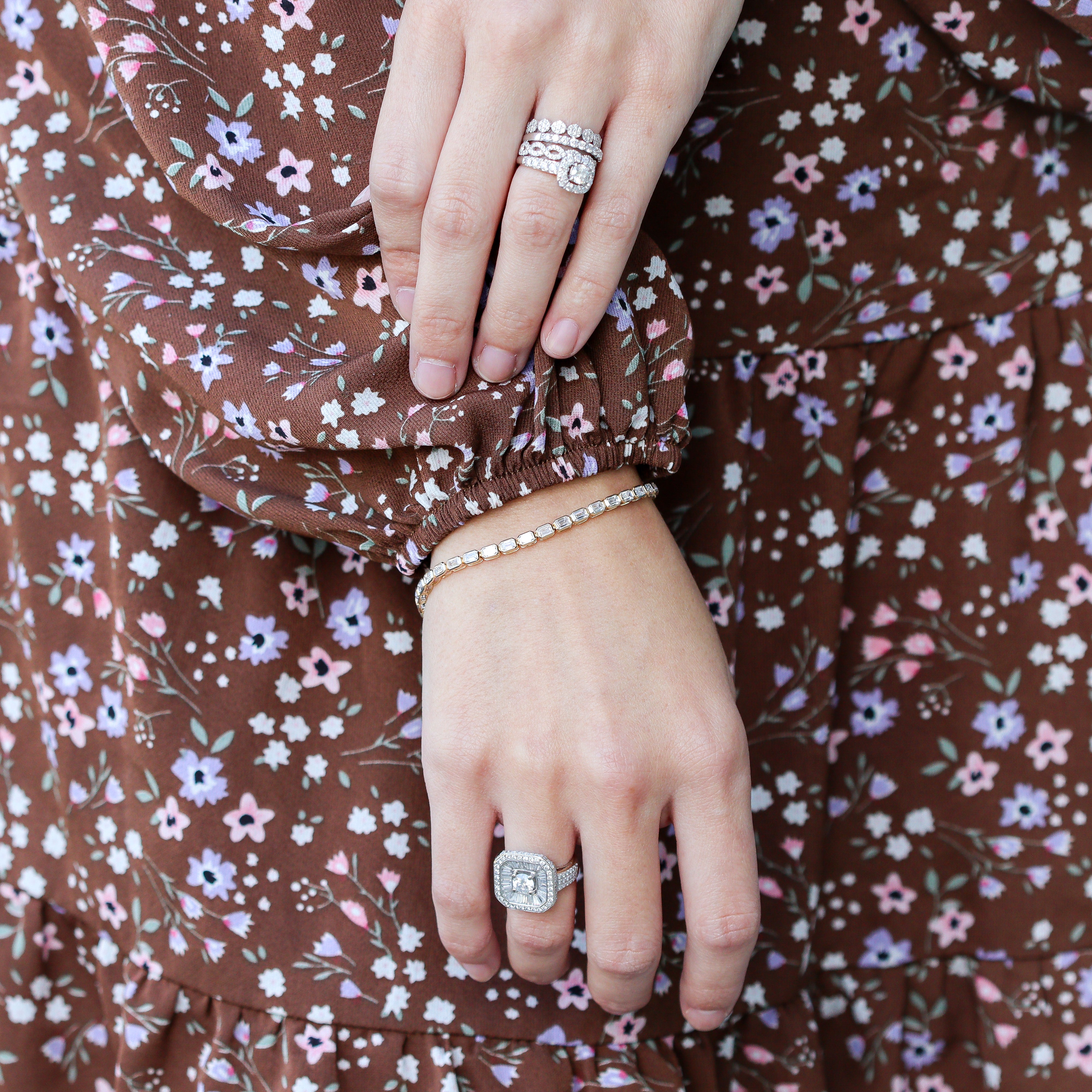 Zoomed out image of tennis bracelet on a model's wrist. She is wearing a floral dress and white gold diamond rings on her hand.