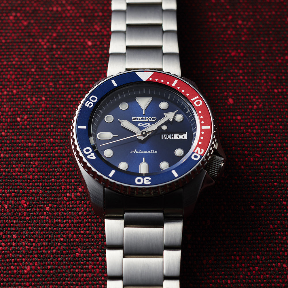 Seiko 5 Sports SRPD53 watch on red-and-black fabric, showing the blue dial, Pepsi bezel and brushed stainless steel bracelet.