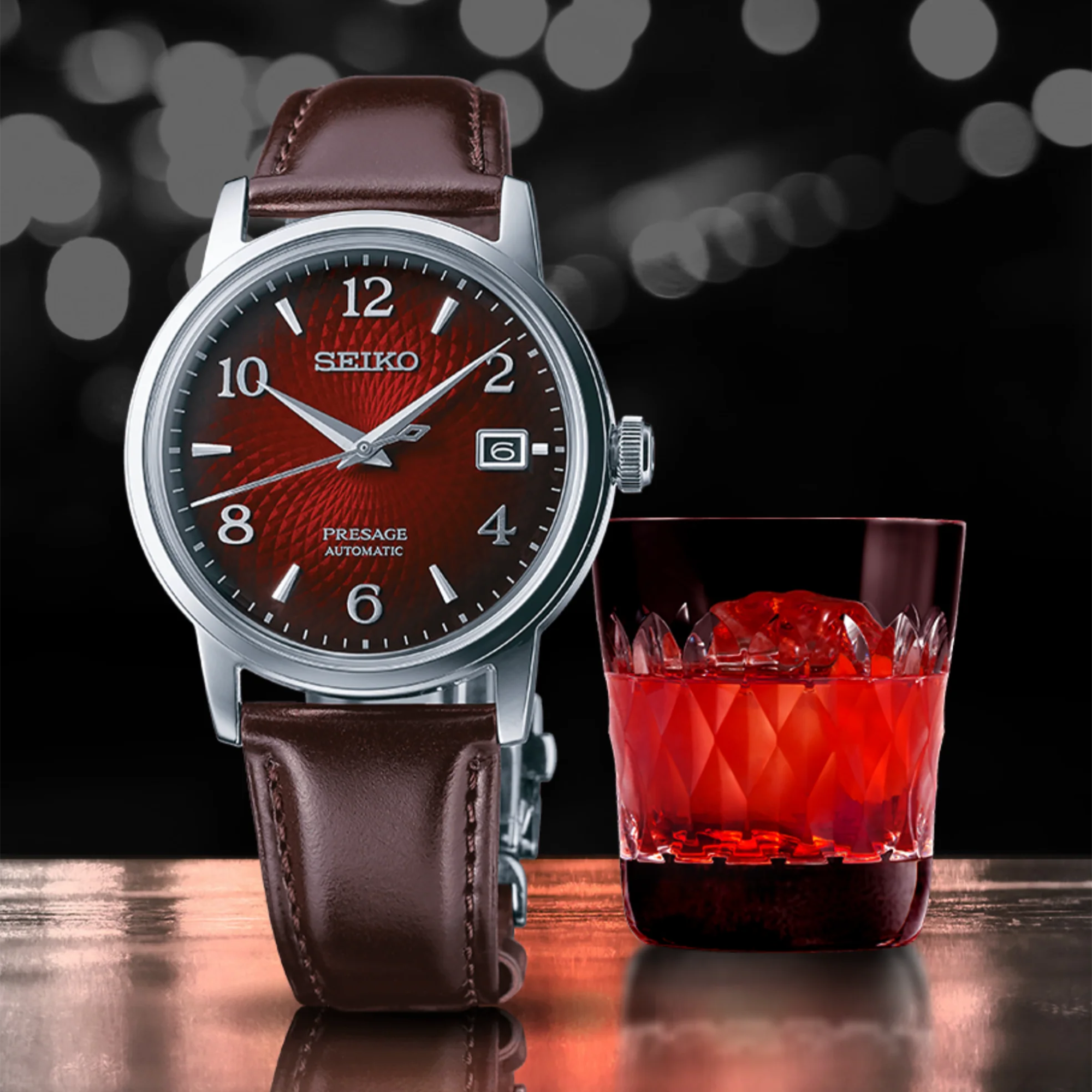 Seiko watch with brown leather strap next to a glass of red liquid on a dark background