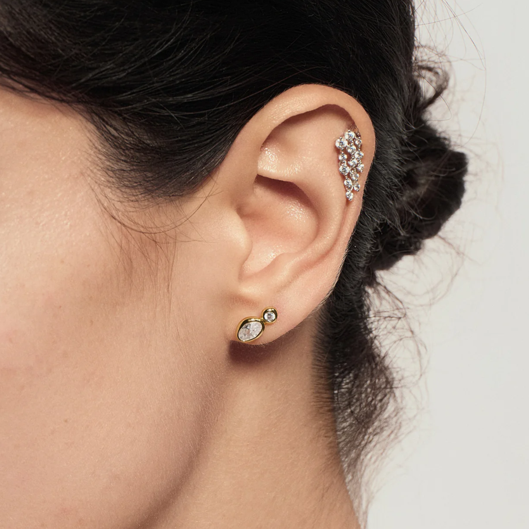 Close-up of a model's ear wearing a gold ear cuff with a cluster of clear stones and a small gold stud earring with a clear oval stone.