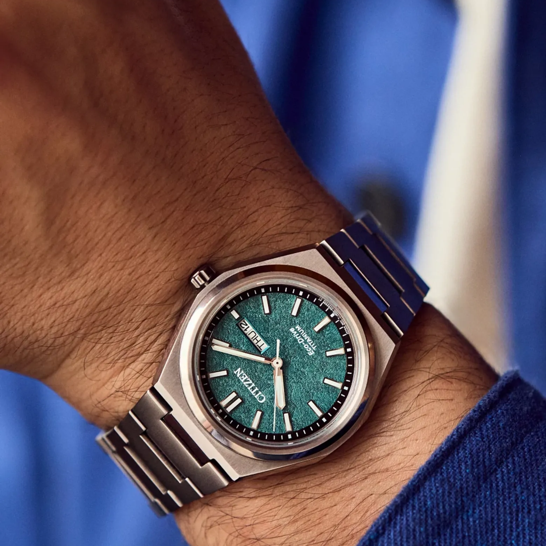 Wrist wearing a watch with a blue strap and green dial against a blurred background