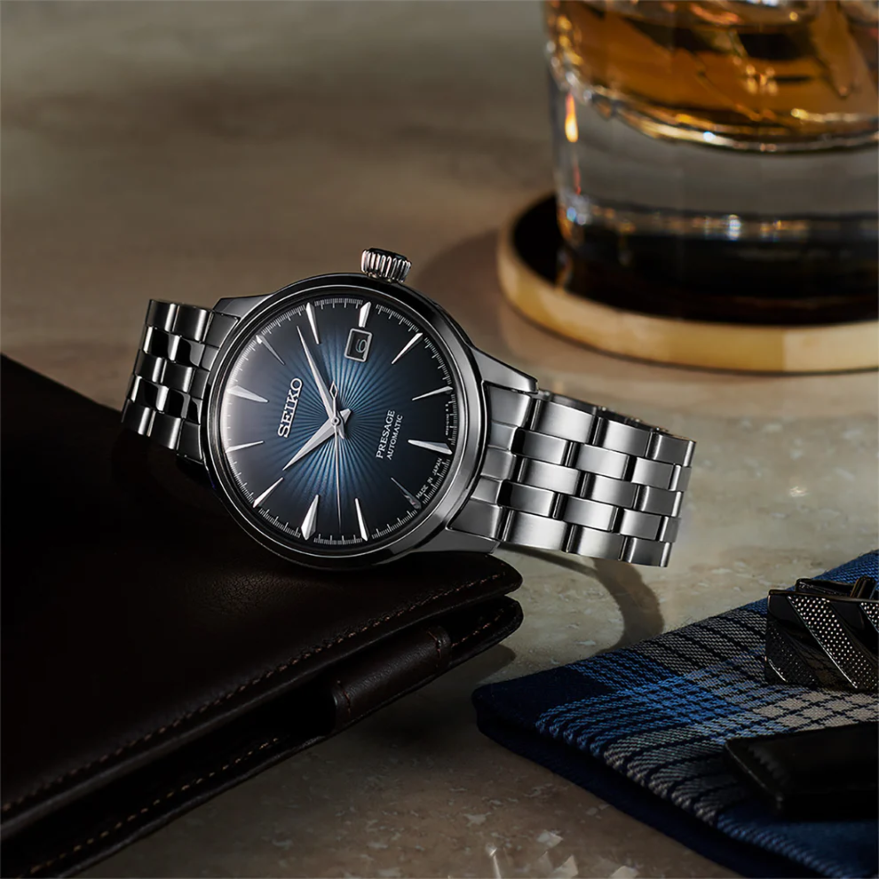 Silver Seiko watch on a marble surface with a glass of whiskey in the background