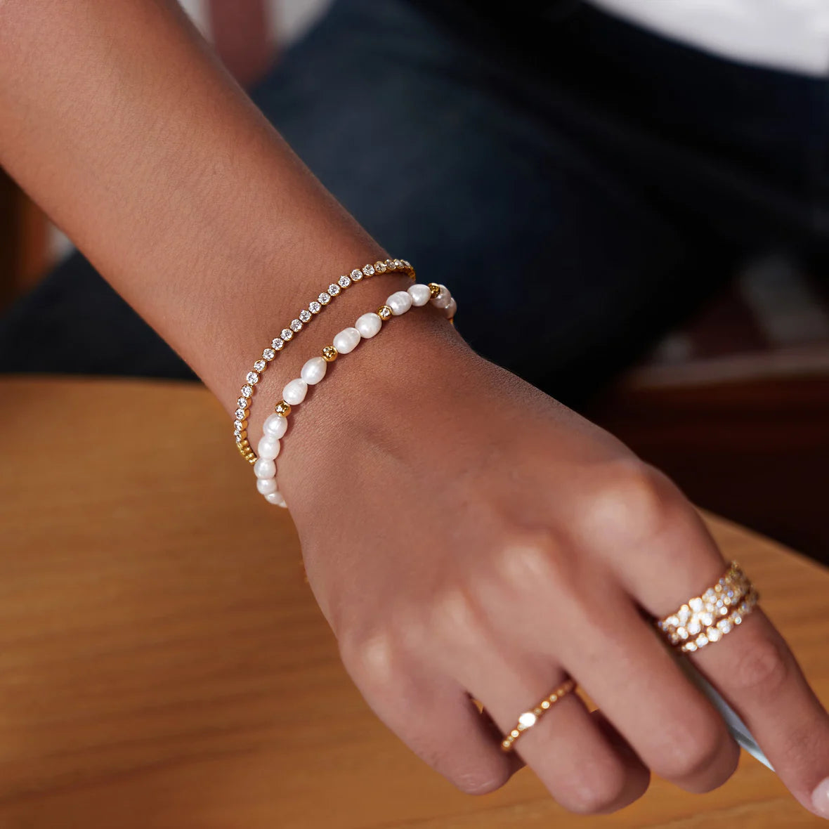 Hand wearing two pearl bracelets and a ring on a wooden surface