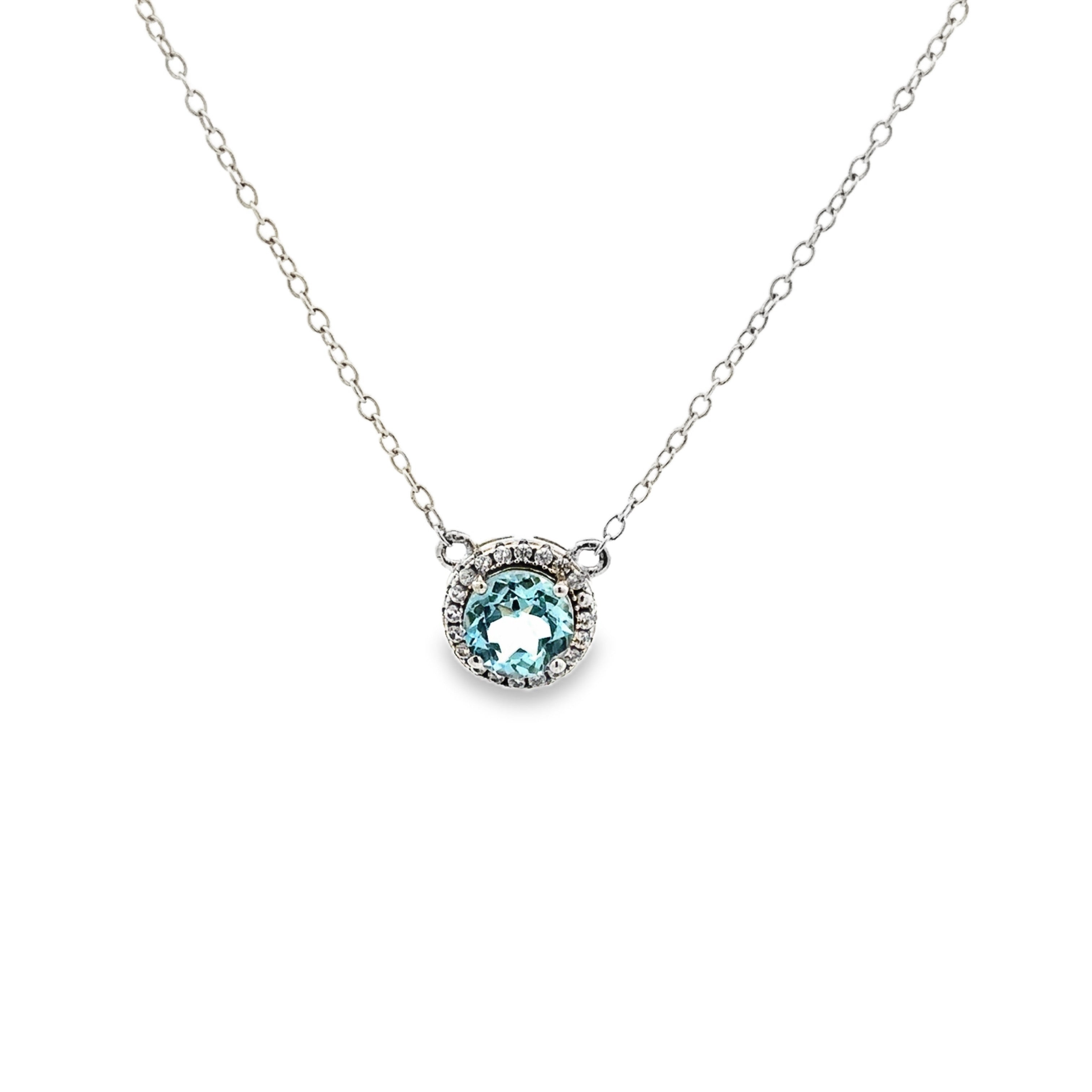 A silver chain necklace with a round pendant featuring a light blue gemstone surrounded by a halo of small clear stones, set against a plain white background.
