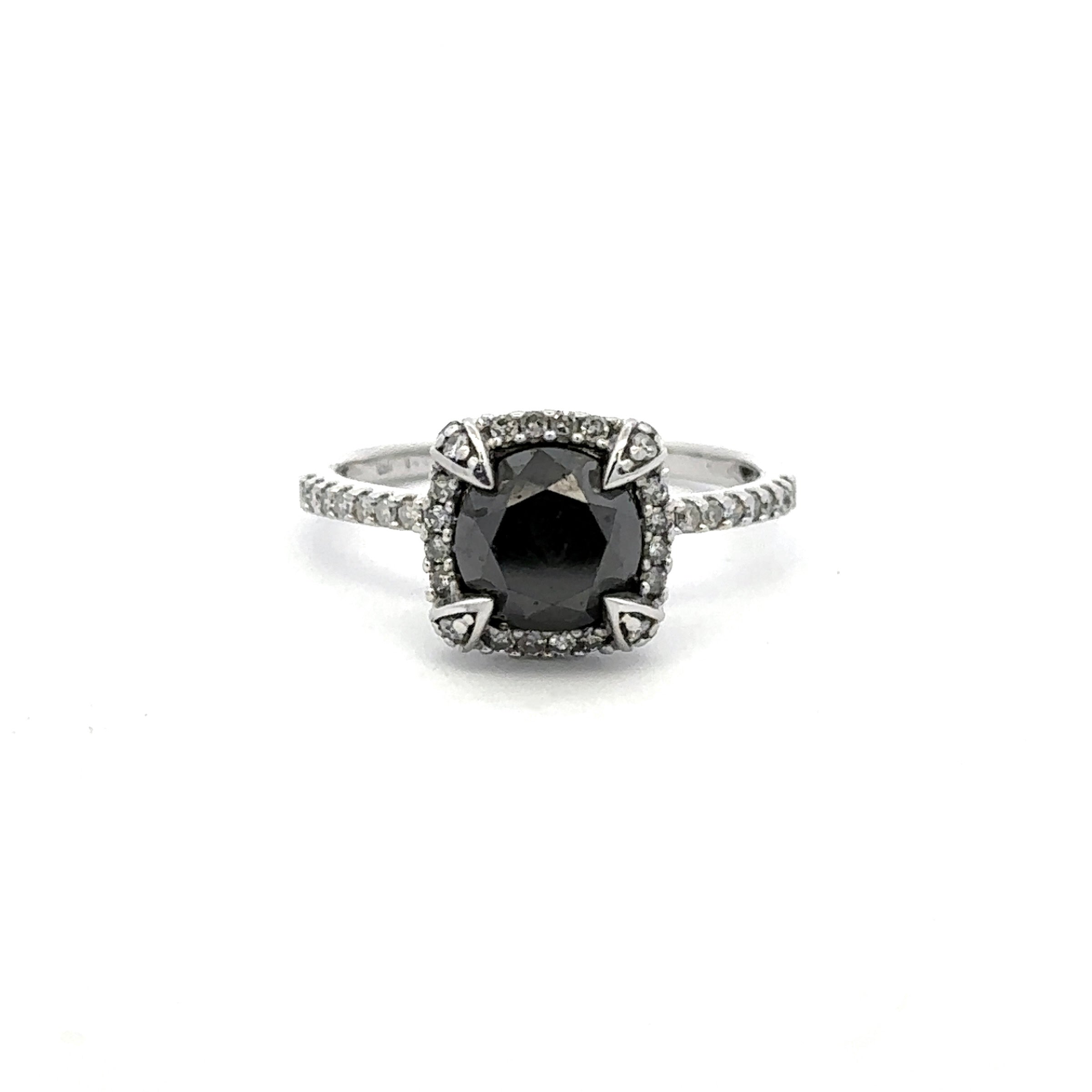 A silver-toned ring featuring a cushion-cut black gemstone surrounded by a halo of small clear stones, with a pavé band, set against a plain white background.
