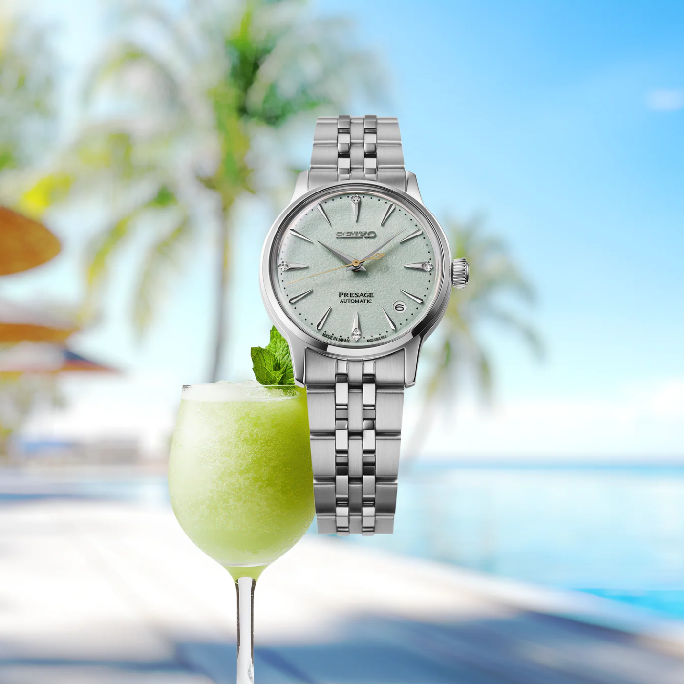 Silver Seiko watch next to a glass of green drink with a blurred outdoor background