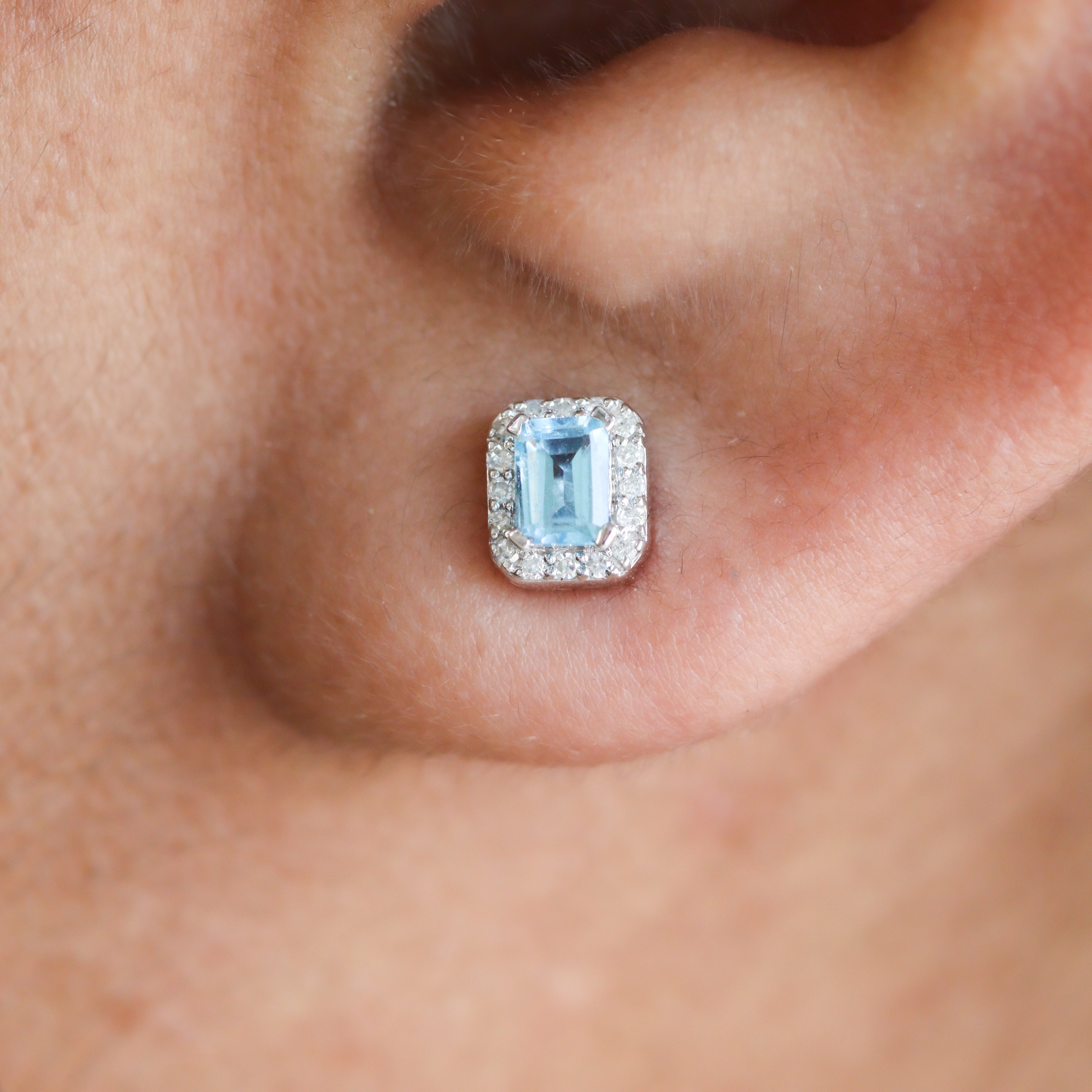 Closeup of 10K White Gold Emerald-Cut Swiss Blue Topaz & 1/10CT. Diamond Halo Stud Earrings on a model's ear.