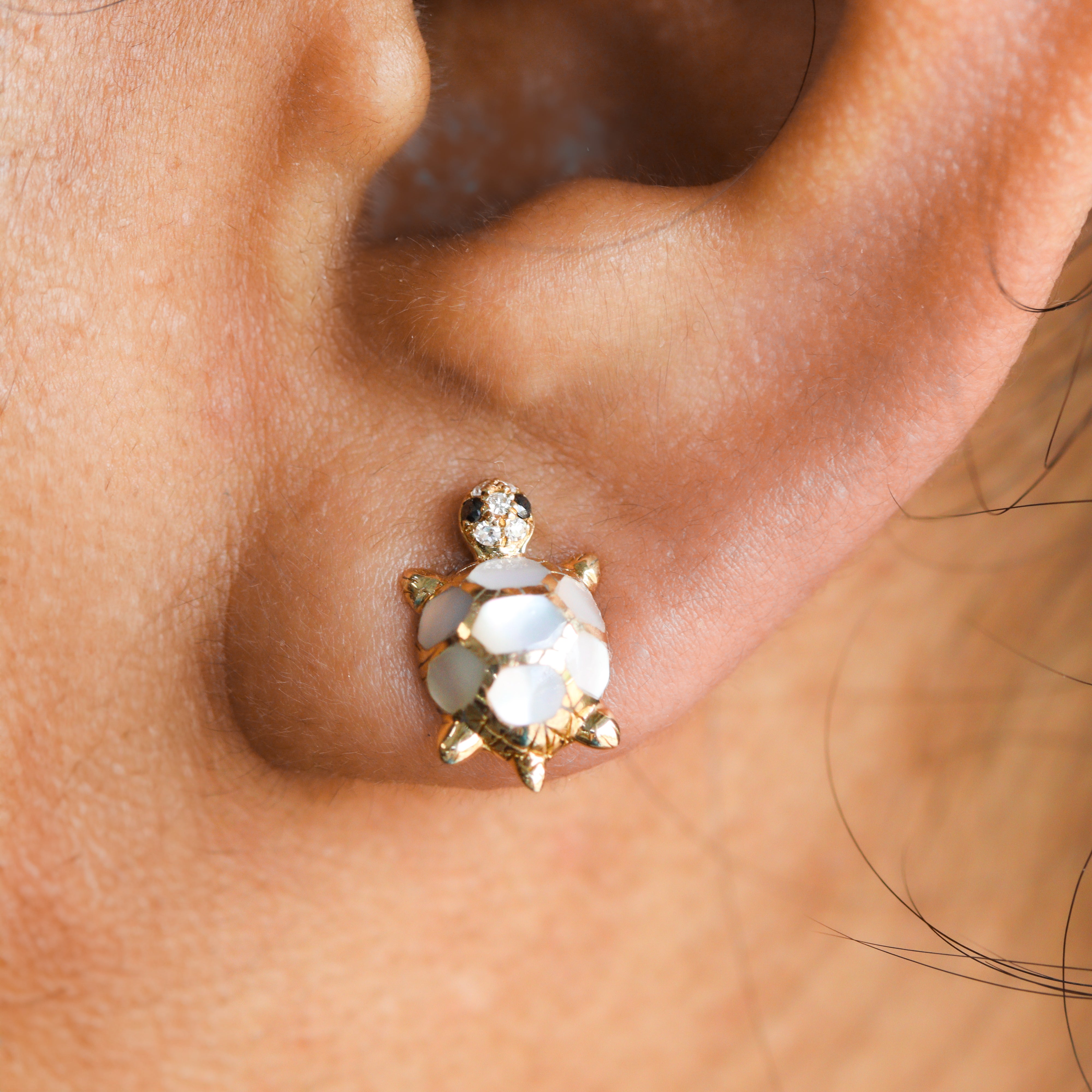 A close up of one 14K Yellow Gold turtle stud earring with mother of pearl in the shell and diamonds in the head, on a model's ear.
