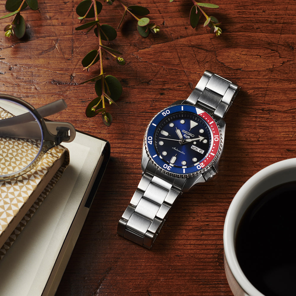 Seiko 5 Sports SRPD53 stainless steel watch on a wooden tabletop beside books, eyeglasses, greenery and a cup of coffee.