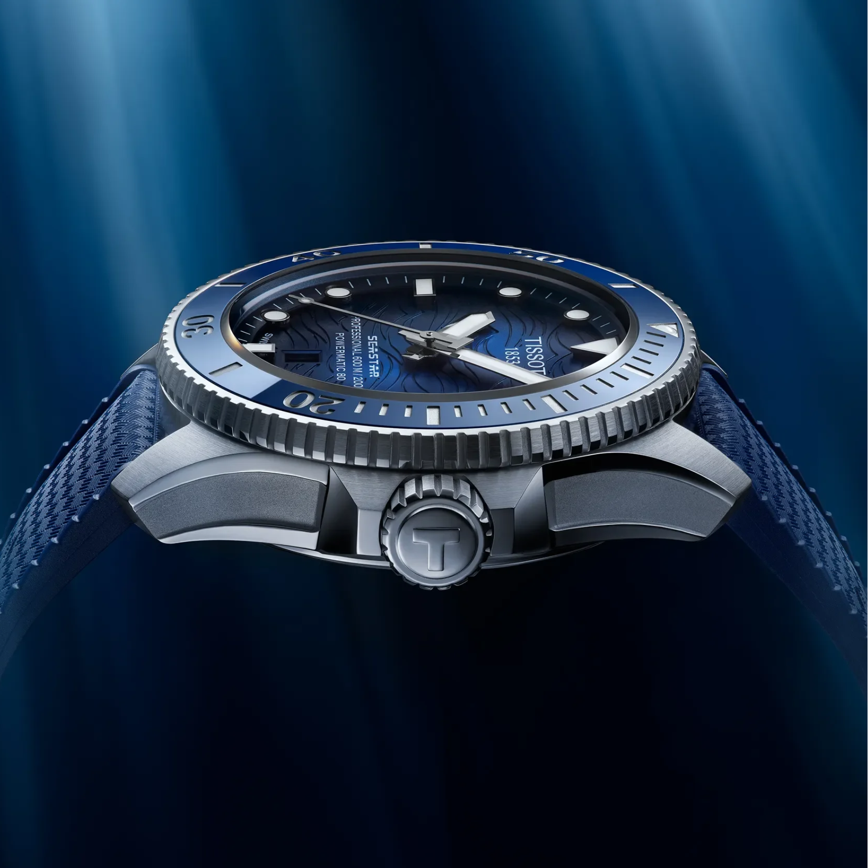 A Tissot diver's watch with a blue dial, blue bezel, and textured blue strap, shown in a close-up side profile against a dark blue background with light rays.