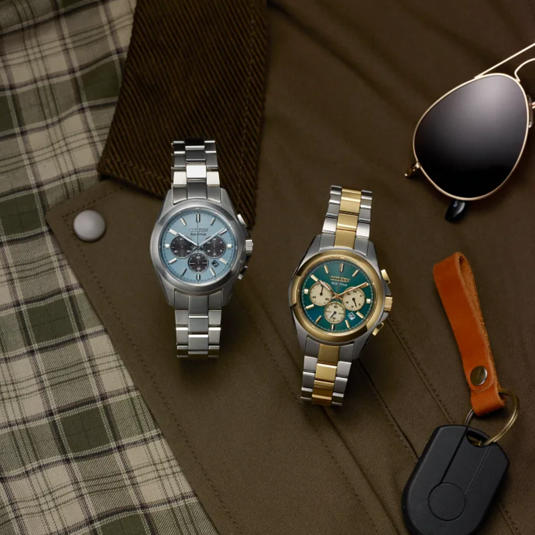 Two watches on a brown surface with plaid fabric and sunglasses.