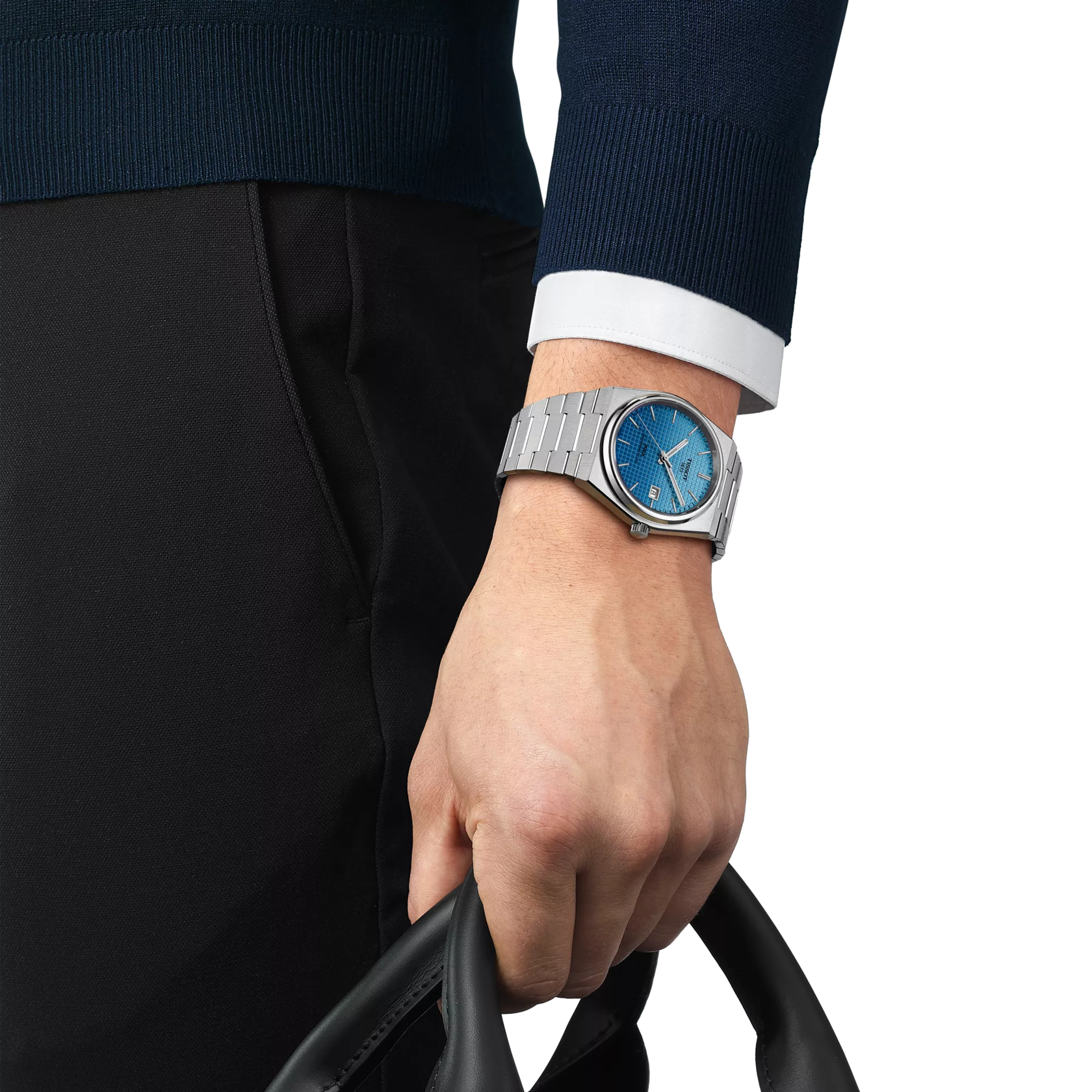Hand wearing a silver watch with a blue face, holding a black bag against a white background.