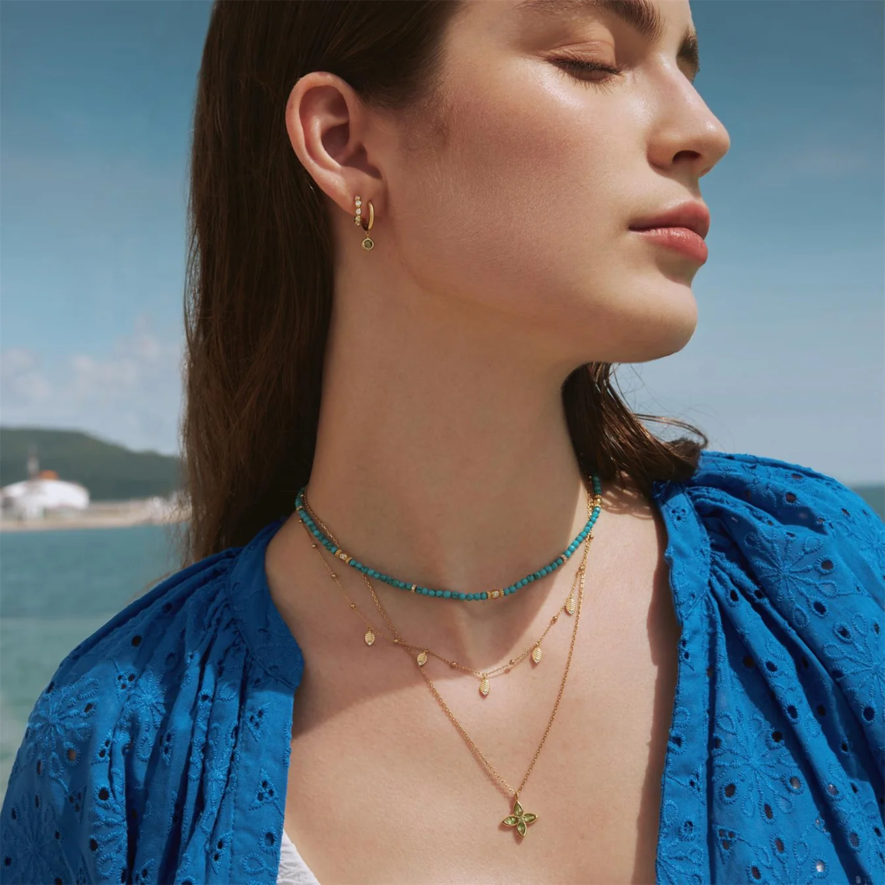 Woman wearing gold and green necklaces with a blue top against a scenic background