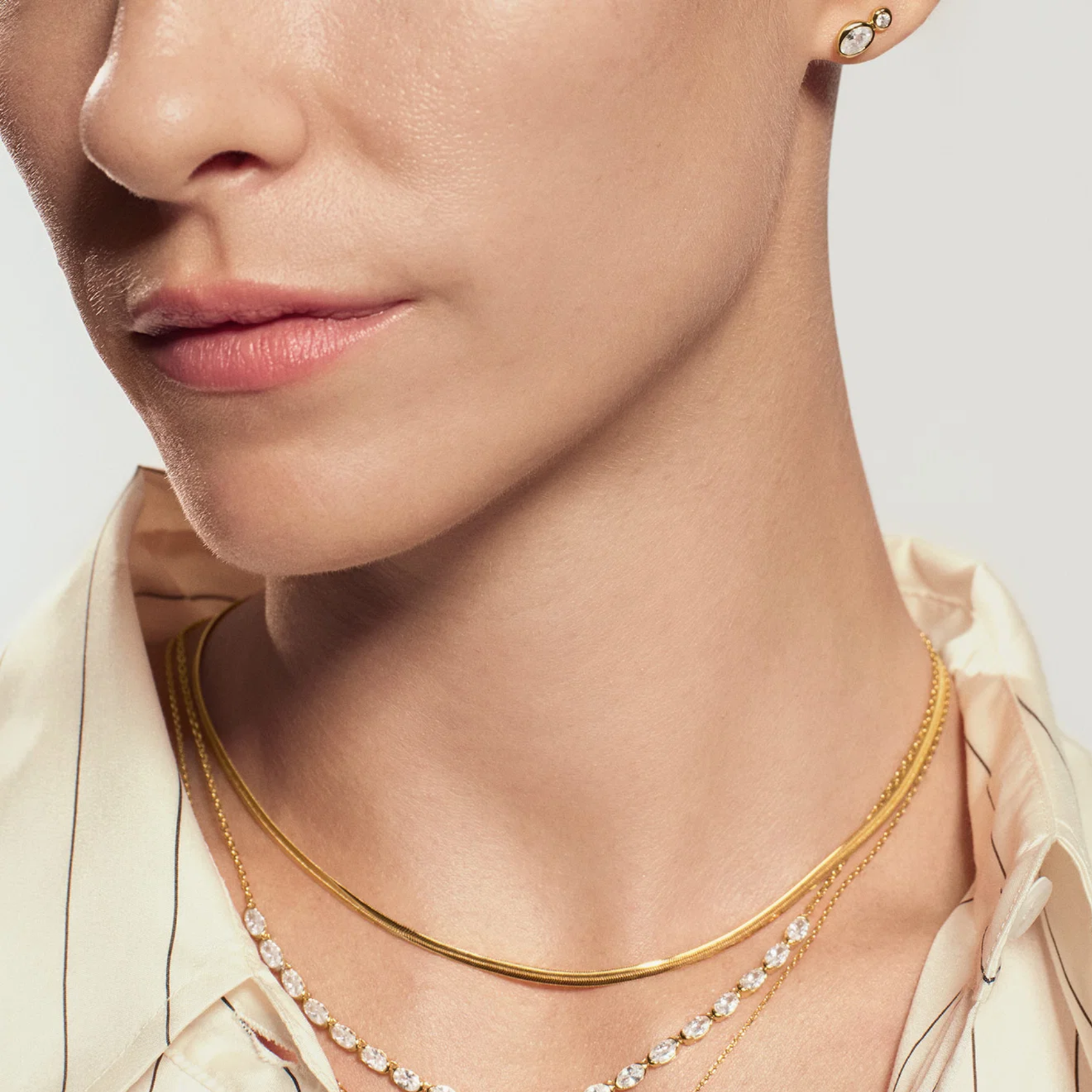 Close-up of a model wearing layered gold necklaces, including a thin chain necklace and a delicate chain with small clear stones, paired with a cream-colored pinstriped shirt.