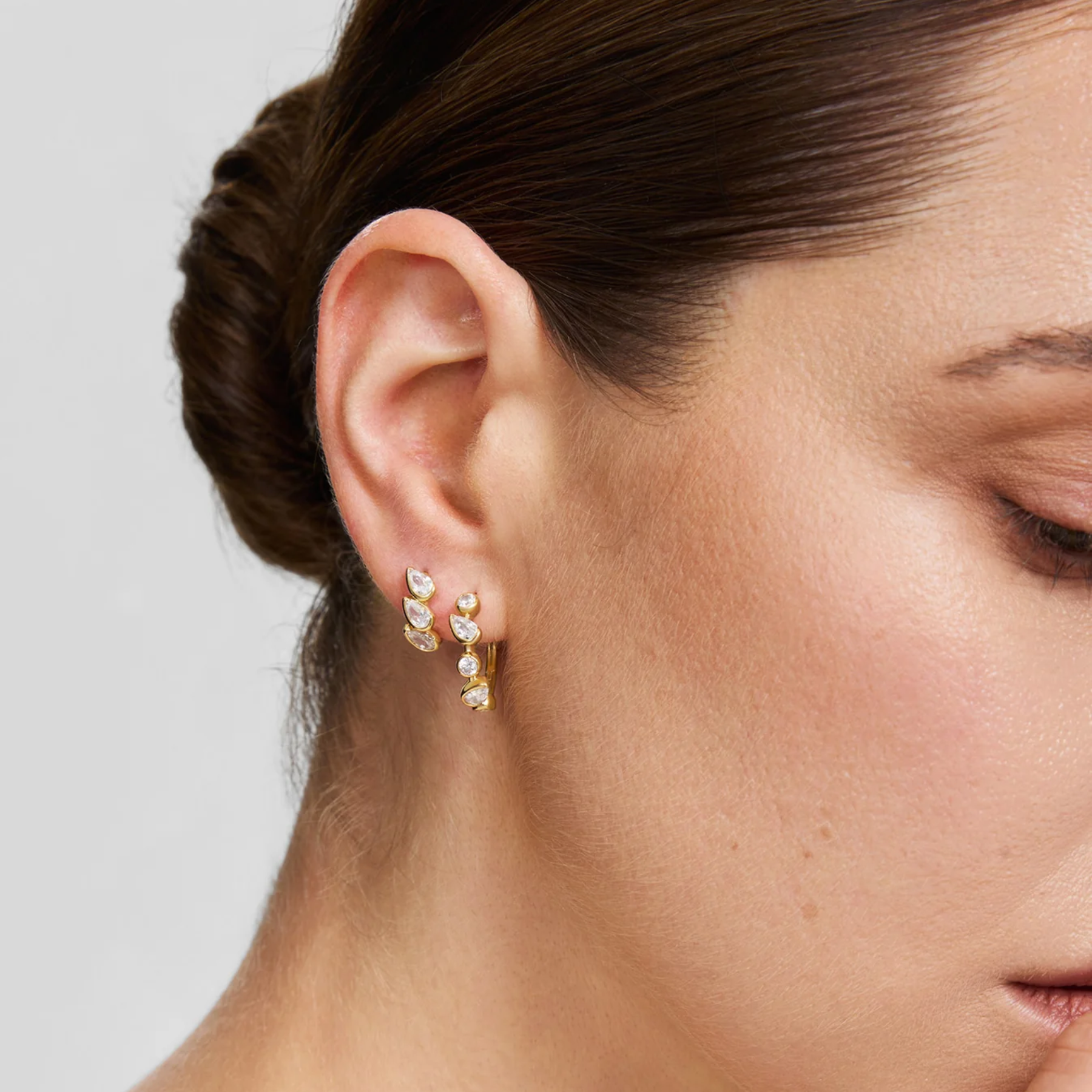 A close-up of a woman's ear wearing a small gold hoop earring with three clear teardrop-shaped crystals, set against a neutral light gray background.