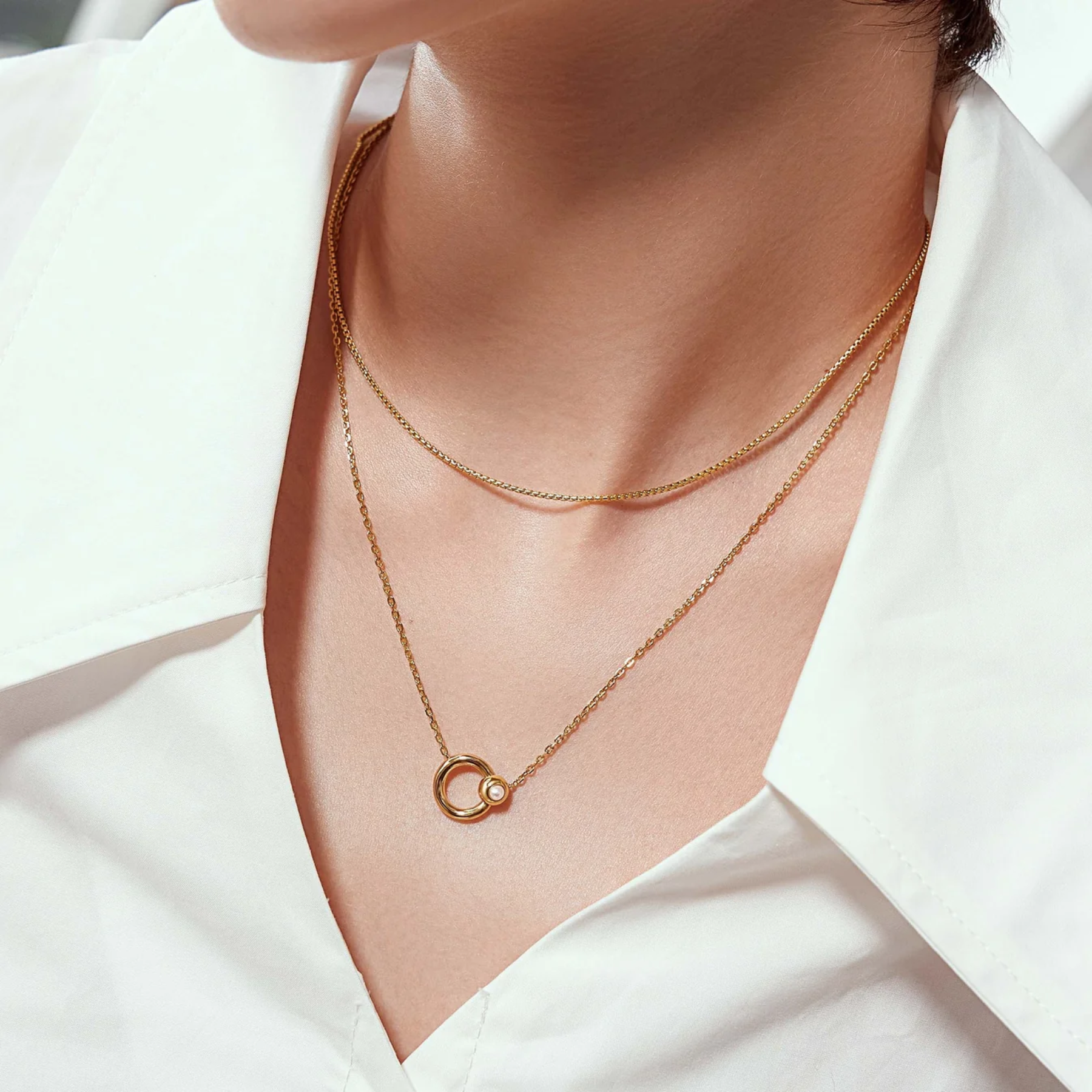A close-up of a person's neck and upper chest wearing two delicate gold necklaces: a fine chain necklace with a small interlocking ring pendant and a slightly thicker chain necklace with a small circular bead accent.