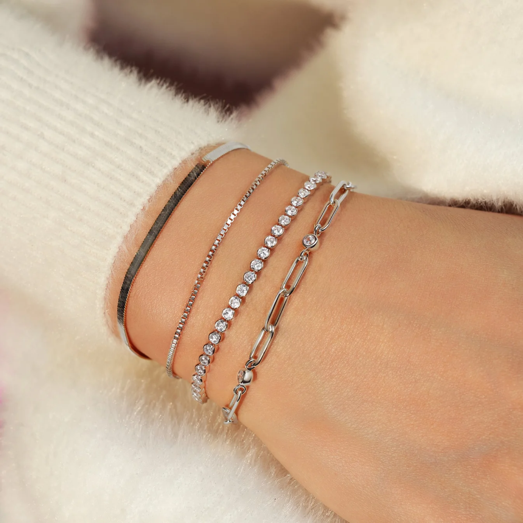 A close-up of a person's wrist wearing four silver-toned bracelets, including a bangle, a beaded chain, a clear-stone link bracelet, and an oval-link chain, styled with a cream knit sleeve against a soft white background.