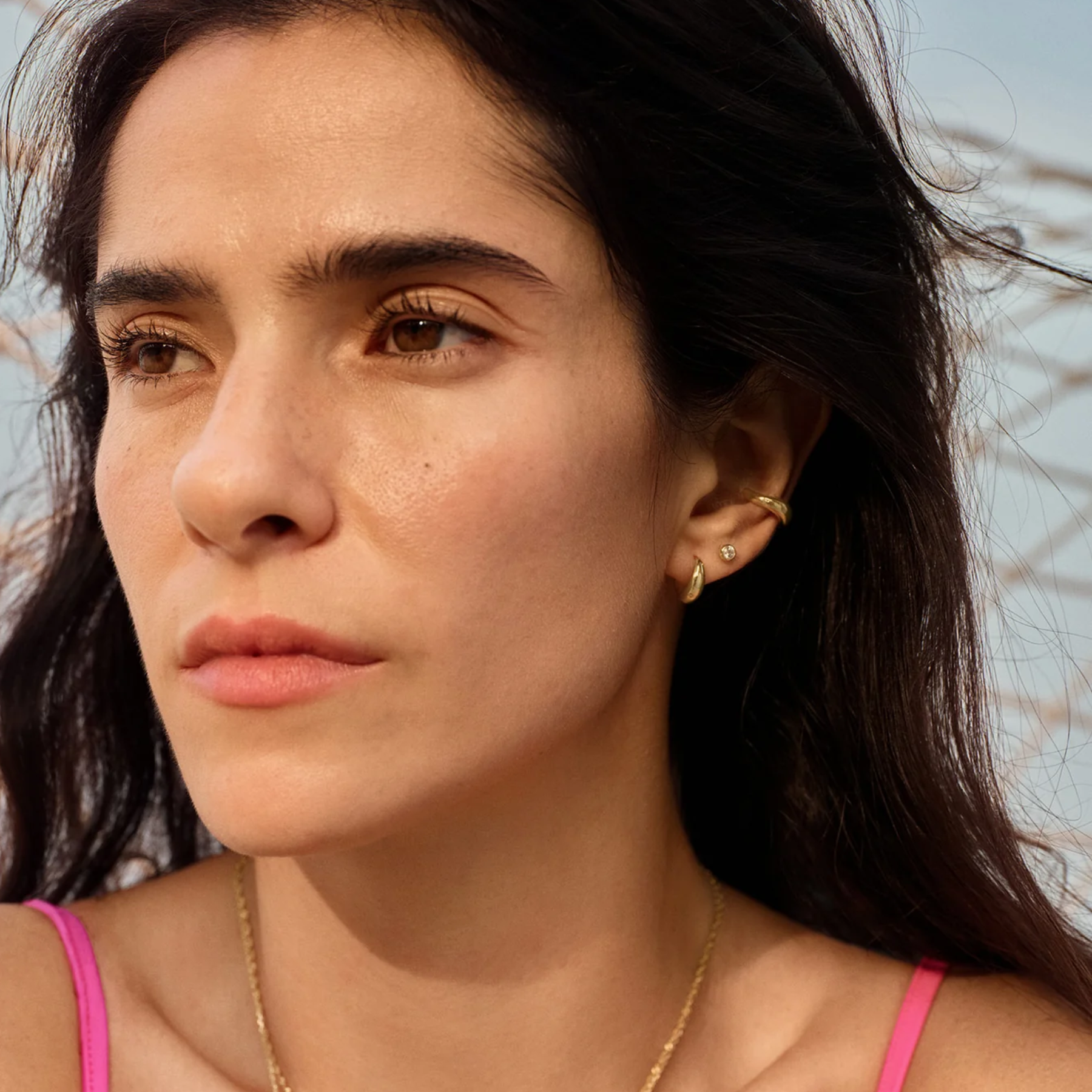 A close-up portrait of a woman with dark hair wearing delicate gold earrings and a gold chain necklace, with a pink top strap visible against a soft, blurred outdoor background.