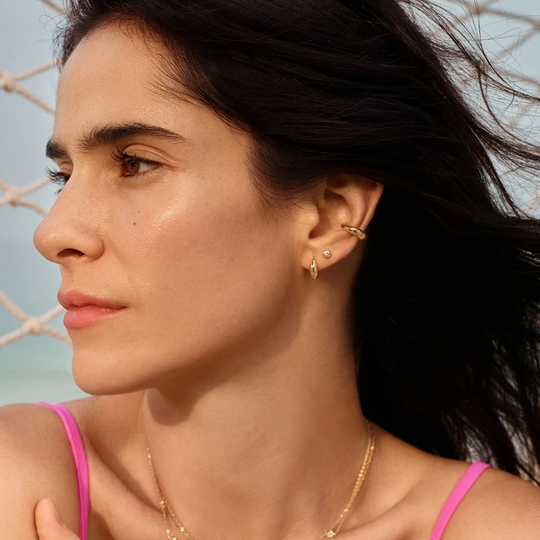 A close-up side profile of a woman wearing gold ear cuff and ear stud jewelry, with a pink bikini top visible against a soft beach-like background.